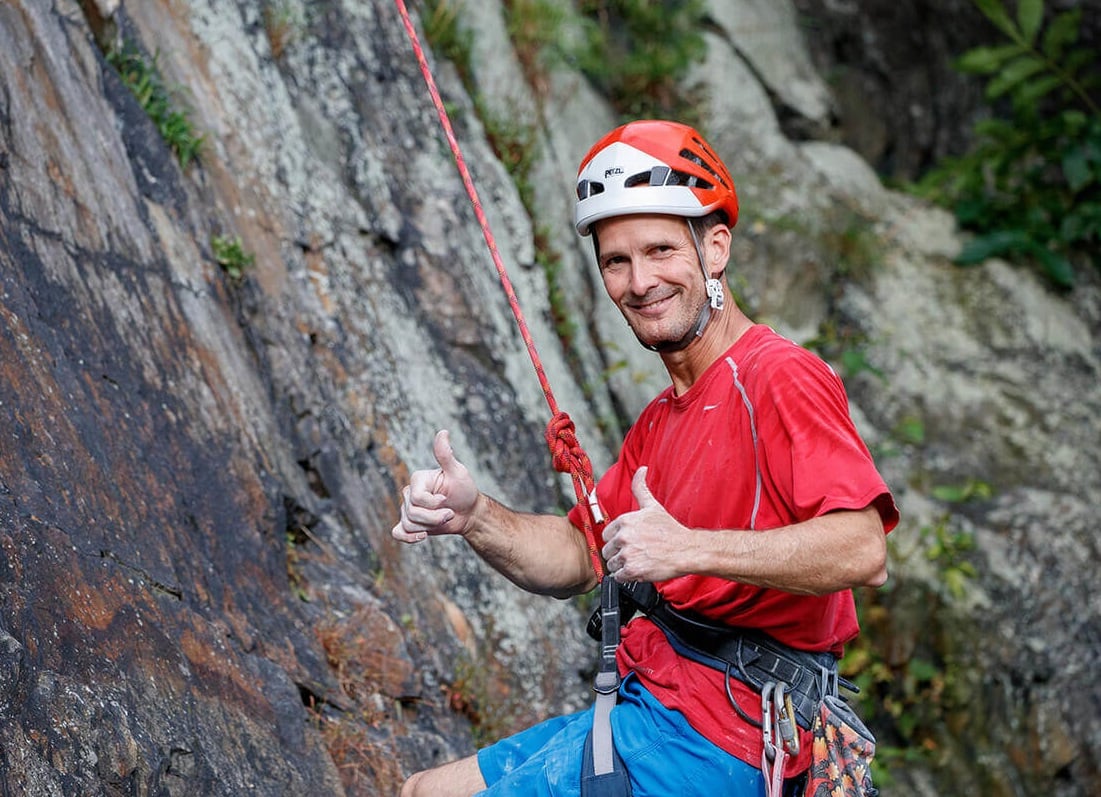 Man in red shirt wearing climbing gear giving thumbs up sign.