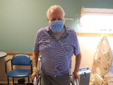 Gabriel standing with the aid of a walker in his hospital room wearing a mask.