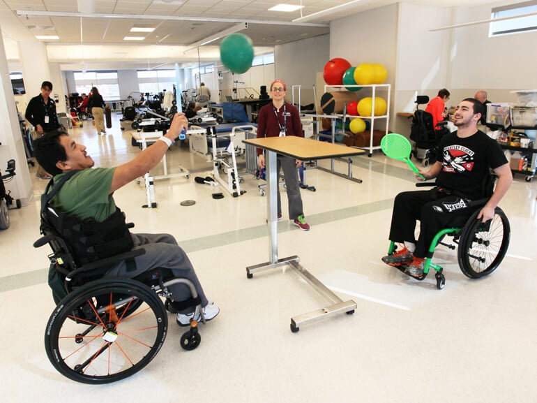 Two male patients in wheelchairs working on coordination skills using ball and paddle.