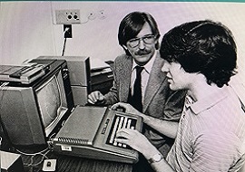Two men sitting at a desk typing into a keyboard connected to small TV monitor.
