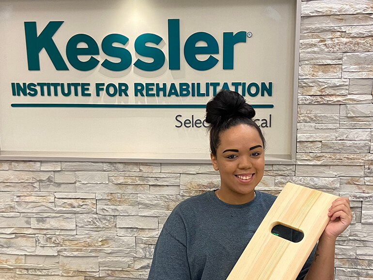 Jasmine standing in front of a Kessler logo sign in the hospital holding a wooden transfer board.