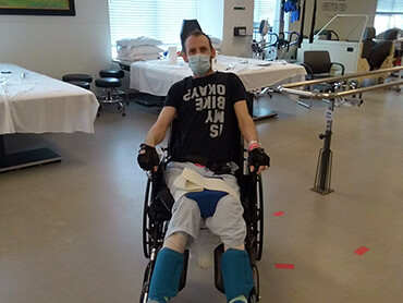 Man wearing a black shirt and hospital mask sitting in a wheelchair in an exercise room.