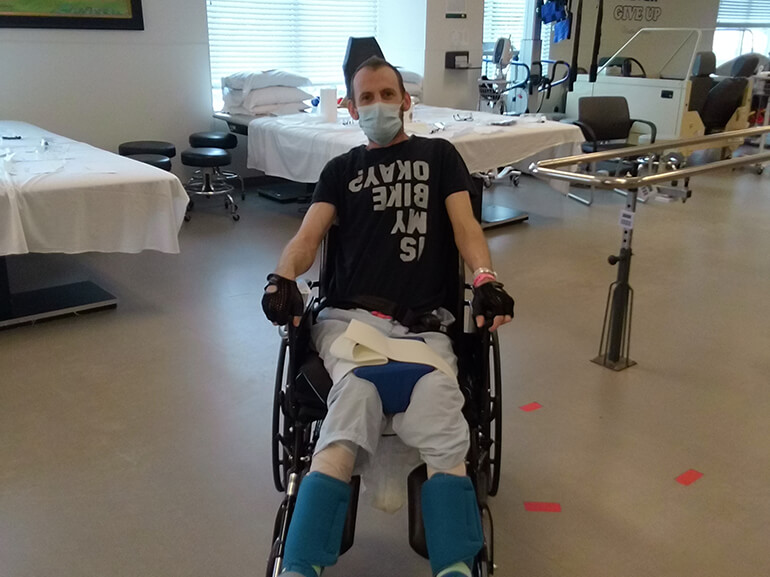 Jason wearing a black t-shirt and hospital mask, sitting in a wheelchair in a therapy gym.