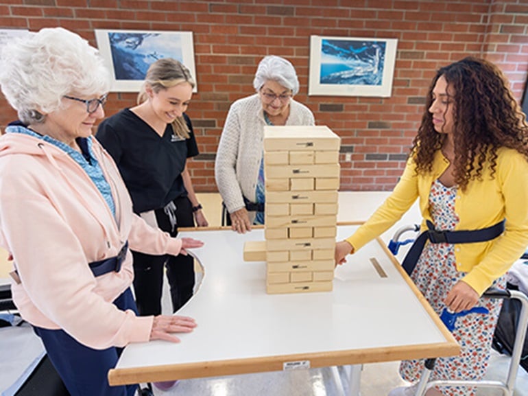 In a rehabilitation space, several people stand around a table and take turns removing blocks from a stacked wooden tower during a group activity.