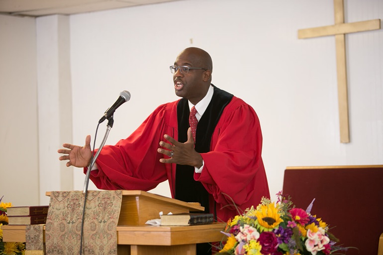 Black man wearing red robe and standing at a small altar preaching.