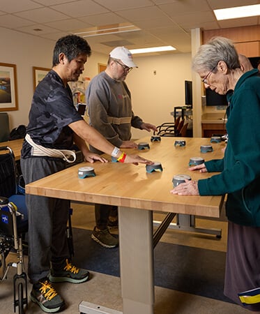 Three stroke patients standing with assistance of a gait belt at a table and reaching light up buttons