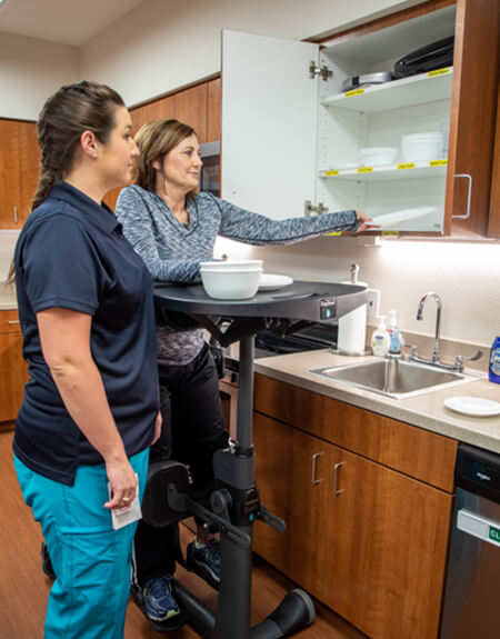 Patient is standing with physical support and reaching for dishes in a cabinet. Occupational therapist is standing beside her