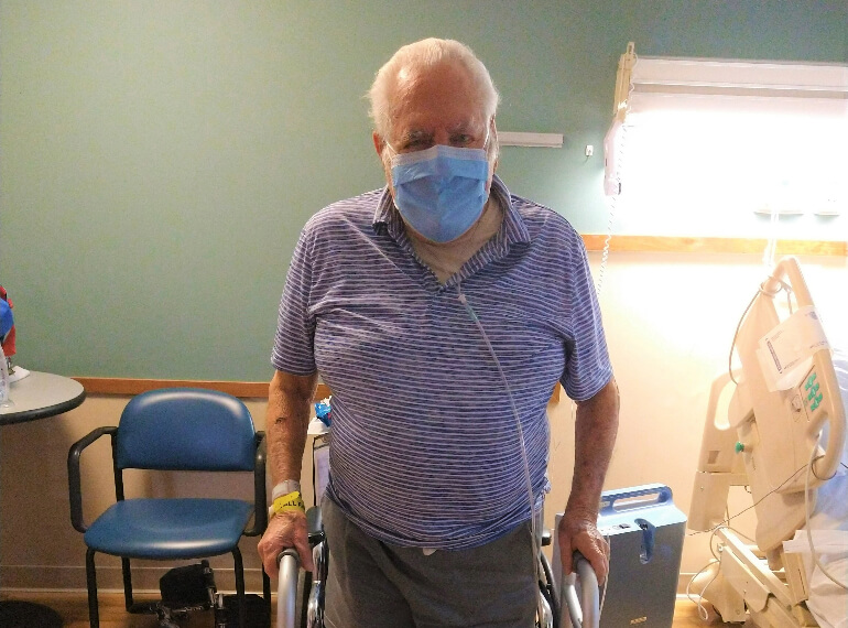 Gabriel standing with the aid of a walker in his hospital room wearing a mask.