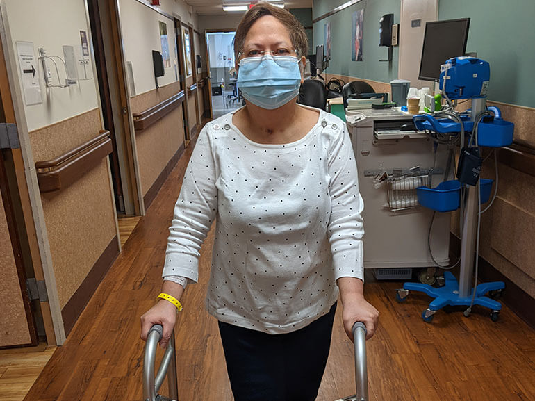 Woman standing outside her hospital room with the aid of a rolling walker wearing a mask.