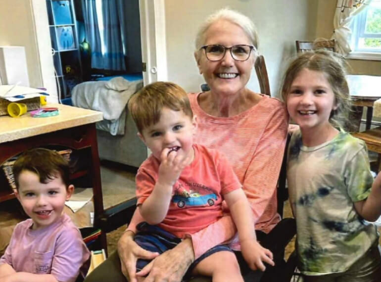 Jane sitting in a wheelchair with her three grandchildren around her.