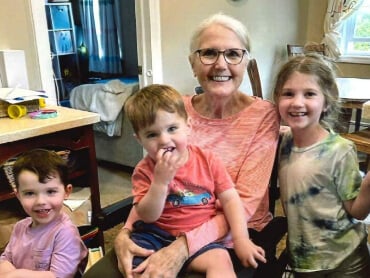 Jane sitting in a wheelchair with her three grandchildren around her.
