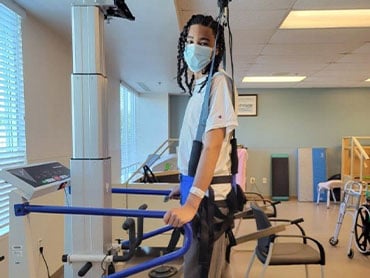 John Parker in the therapy gym walking on the treadmill with the aid of a body weight support harness wearing a mask.