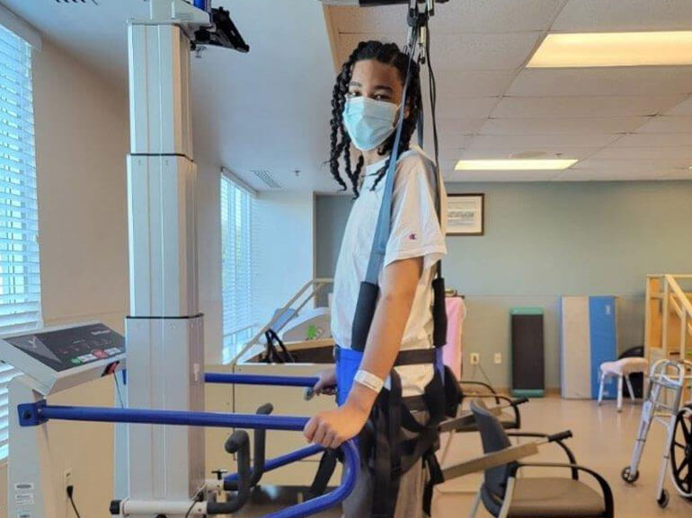 John Parker in the therapy gym walking on the treadmill with the aid of a body weight support harness wearing a mask.