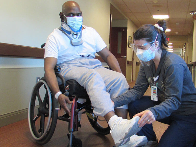 Leroy Pettey in a wheelchair wearing a mask and neck brace  in the hospital hallway with a therapist adjusting his foot.