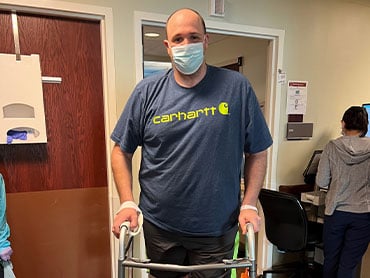 Scott Darcy standing outside his hospital room with the support of a rolling walker.