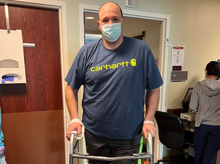 Scott Darcy standing outside his hospital room with the support of a rolling walker.