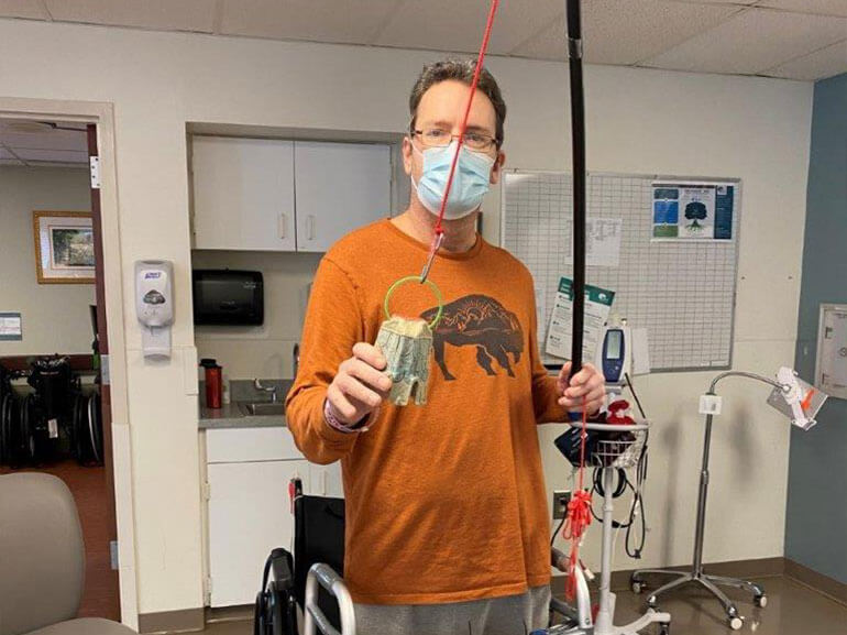 Tom Wolfinger standing in a hospital common area with a mask on holding a fishing pole he used in his occupational therapy.