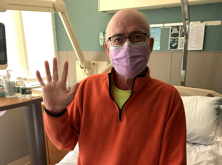 William sitting up on his hospital bed waving and wearing a mask.
